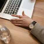A close-up of a woman's watch-adorned hand on a wooden desk beside a laptop and glass of water.